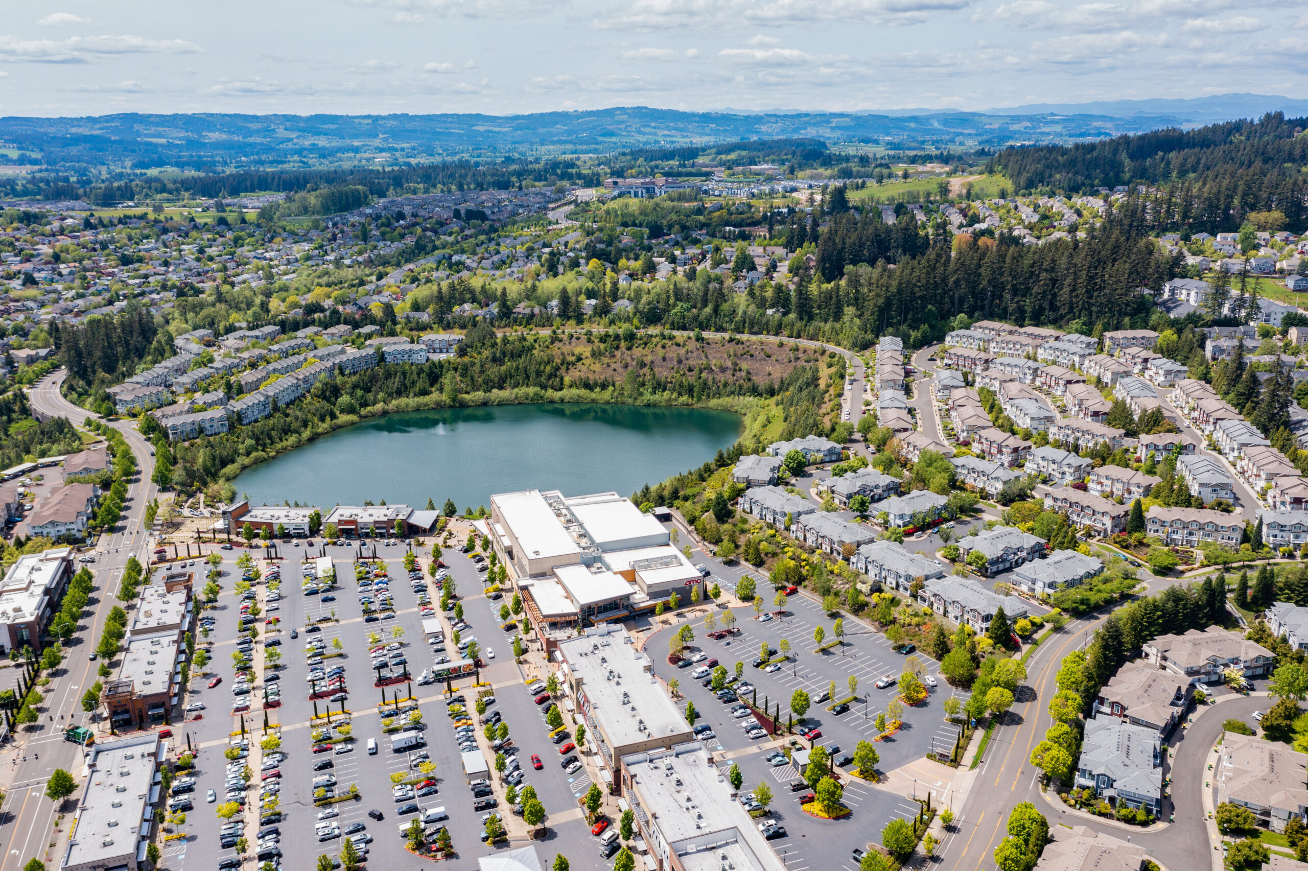 Aerial drone view of Beaverton, Oregon, featuring Progress Lake Park, residential neighborhoods, shopping centers, and parking lots, with tree-lined streets and a mix of suburban and commercial spaces